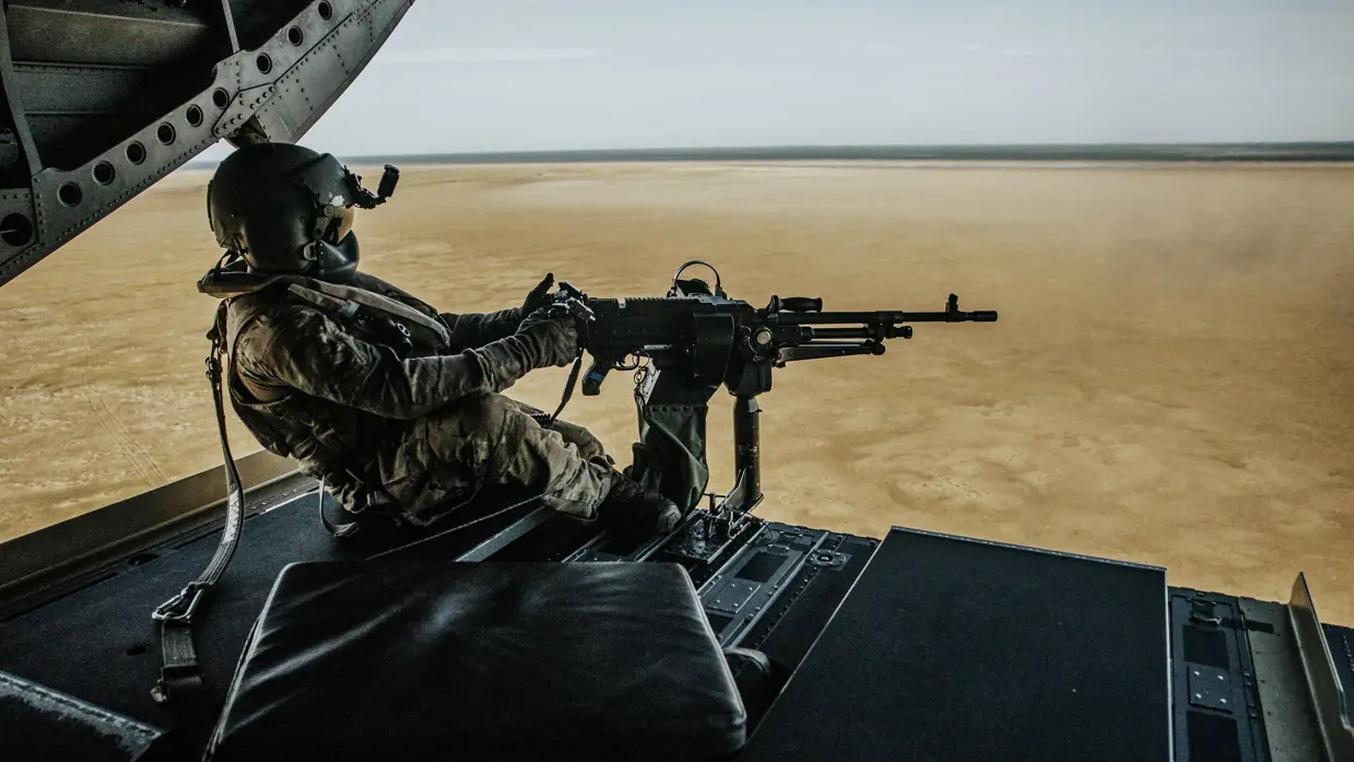 Een militair met een gemonteerd vuurwapen kijkt uit over een landschap vanuit de achterkant van een Chinook-helikopter.