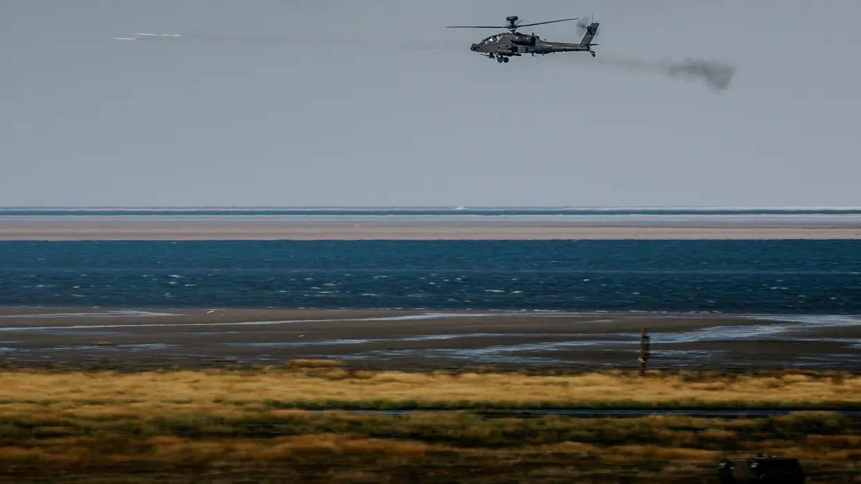 Een landschap met een kustlijn is zichtbaar, met daarboven een vliegende Apache-helikopter die een raket afschiet.
