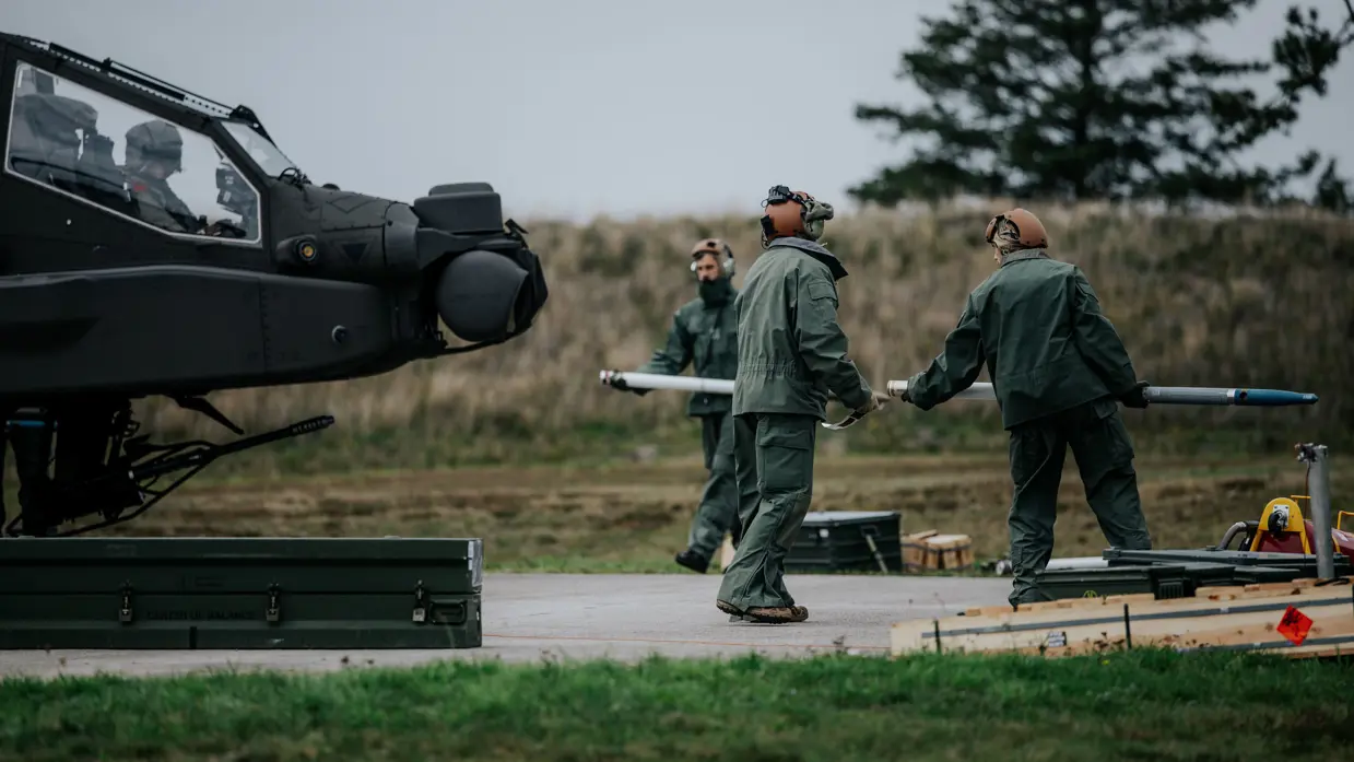 Een drietal militairen tilt raketten op voor een gelande Apache-helikopter.