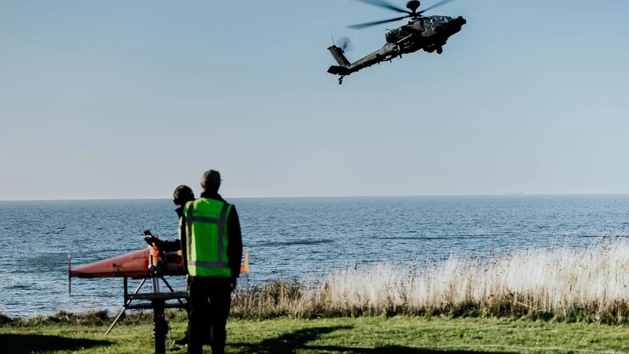 Een Apache-helikopter vliegt boven het water, met een tweetal militairen zichtbaar in de voorgrond.
