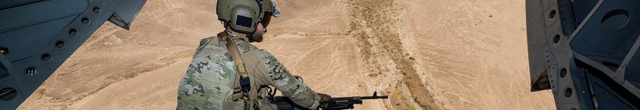 Militair met wapen op ramp van Chinook-transporthelikopter, boven zandlandschap Irak.