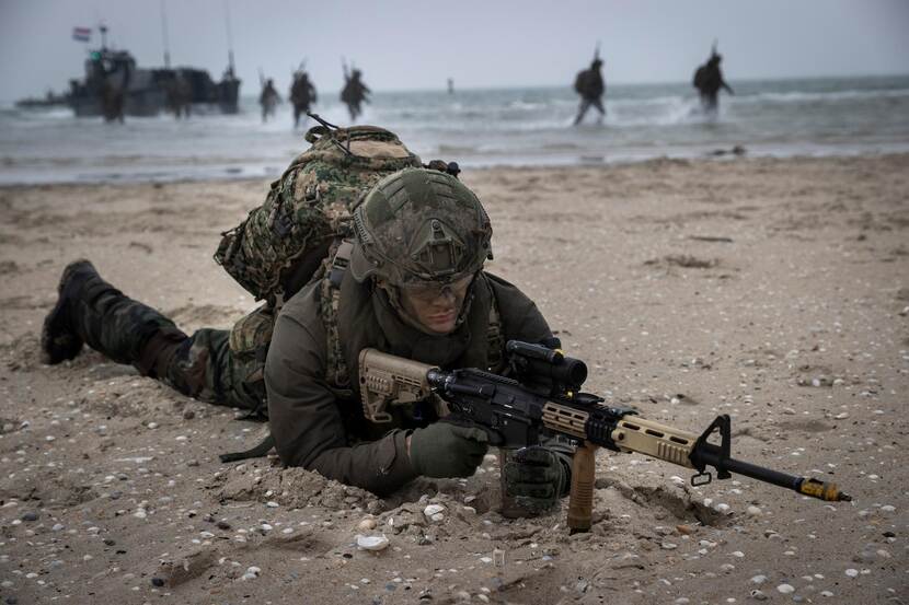 Een militair ligt  met wapen en uitrusting op het strand. Achter hem in zee lopen meer militairen en ligt een vaartuig.