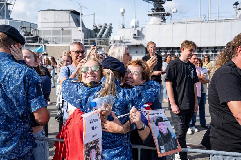 Vrouwelijke militair op de rug gezien wordt door 2 vrouwen in de armen gesloten. Op de achtergrond schip.