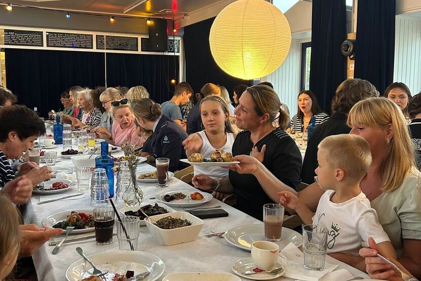 Vrouwen en kinderen eten aan lange tafels en praten met elkaar.