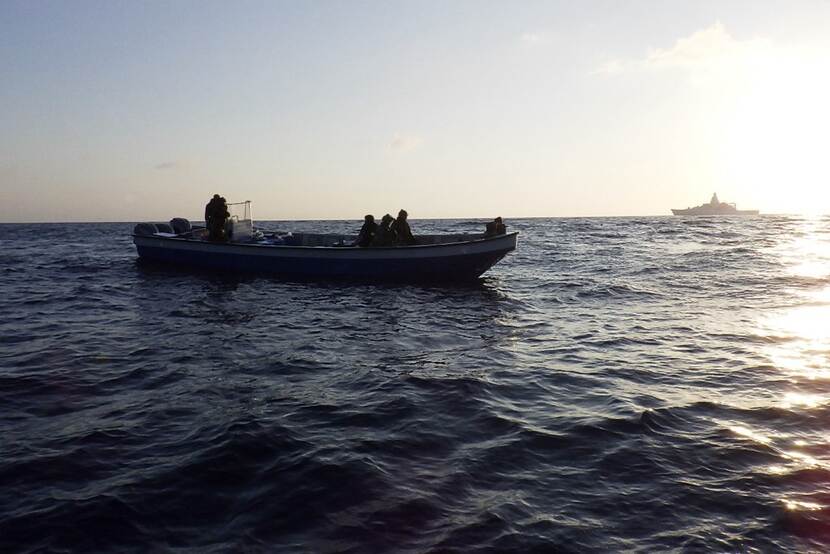 Militairen op onderschept drugsvaartuig, met marineschip op achtergrond.