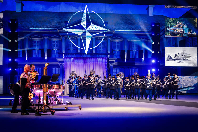 Het Orkest Koninklijke Marechaussee betreedt de vloer. Achter hen op het LED-scherm wordt het NAVO-embleem en foto’s van militair materieel getoond.