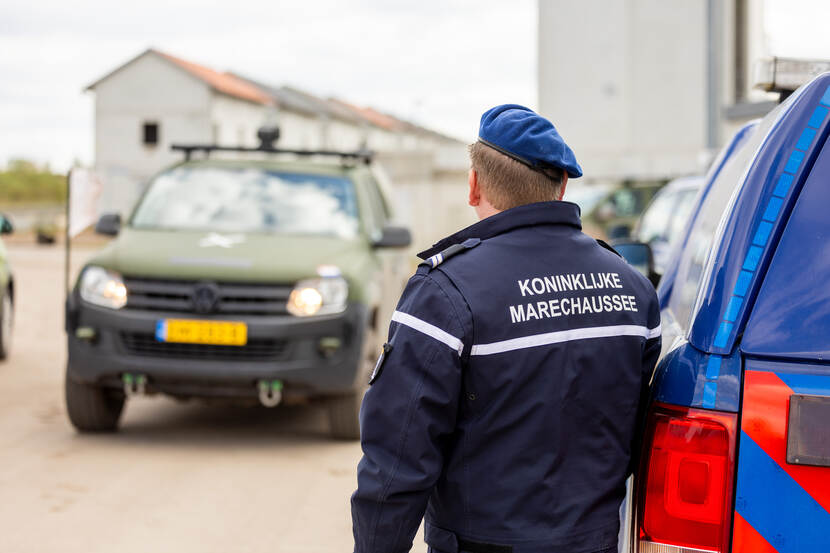 Achterkant van de jas van een marechaussee, staand naast zijn gestreepte auto. Op de achtergrond rijdt een groene Amarok