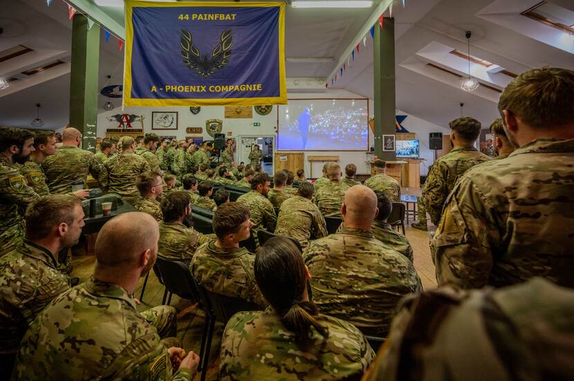 Groep militairen kijkt naar scherm met mensen in een zaal.