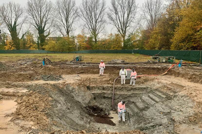 Vier mannen in pakken staan in een diepe kuil. Deze plek in Dordrecht werd een Spitfire geborgen.