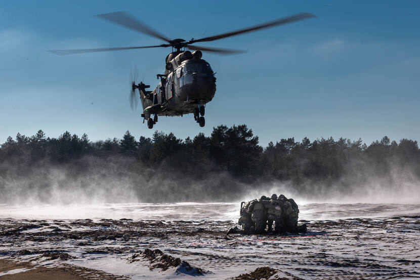 Een Cougar-transporthelikopter landt in een besneeuwd militair oefengebied, de Leusderheide. Een groep militairen wacht op de grond in een kring.