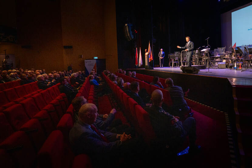 Minister Ruben Brekelmans op het podium, voor hem rode stoelen in het publiek met mensen erop