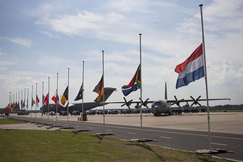 Vlaggen van verschillende nationaliteiten hangen halfstok. Op de achtergrond staan 2 transportvliegtuigen en veel rouwwagens.