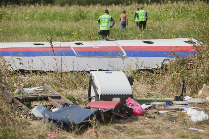 Wrakstukken van een vliegtuig liggen in een veld. Op de achtergrond lopen 4 mensen. 3 met een groen vest en 1 met ontbloot bovenlijf.