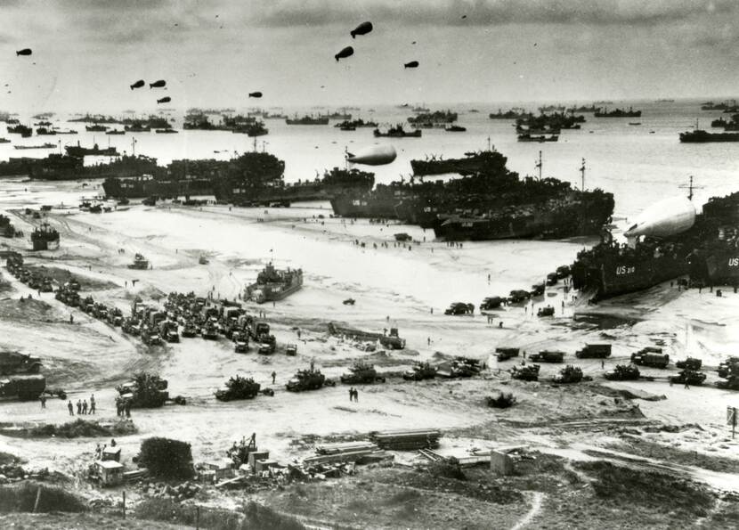 Zwart-witfoto. Een van de invasiestranden in Normandië tijdens D-Day, van bovenaf gezien. Een grote stroom voertuigen verlaat de landingsvaartuigen.