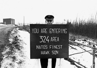 Militair langs weg voor bord met Engelstalige tekst: you are entering 324 area natos finest hawk sqn.