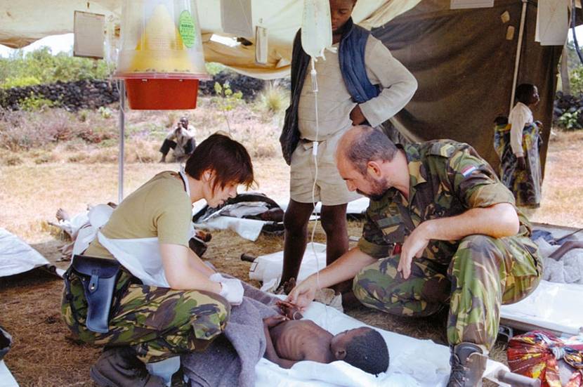 2 militairen van een medisch team verzorgen een vluchteling die met een infuus op een brancard ligt in een vluchtelingenkamp.